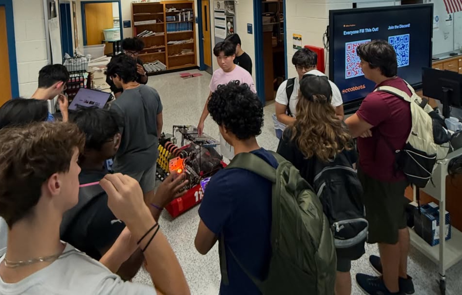 Students gathered around a competition robot, with sign-up and Discord QR codes on the screen behind them